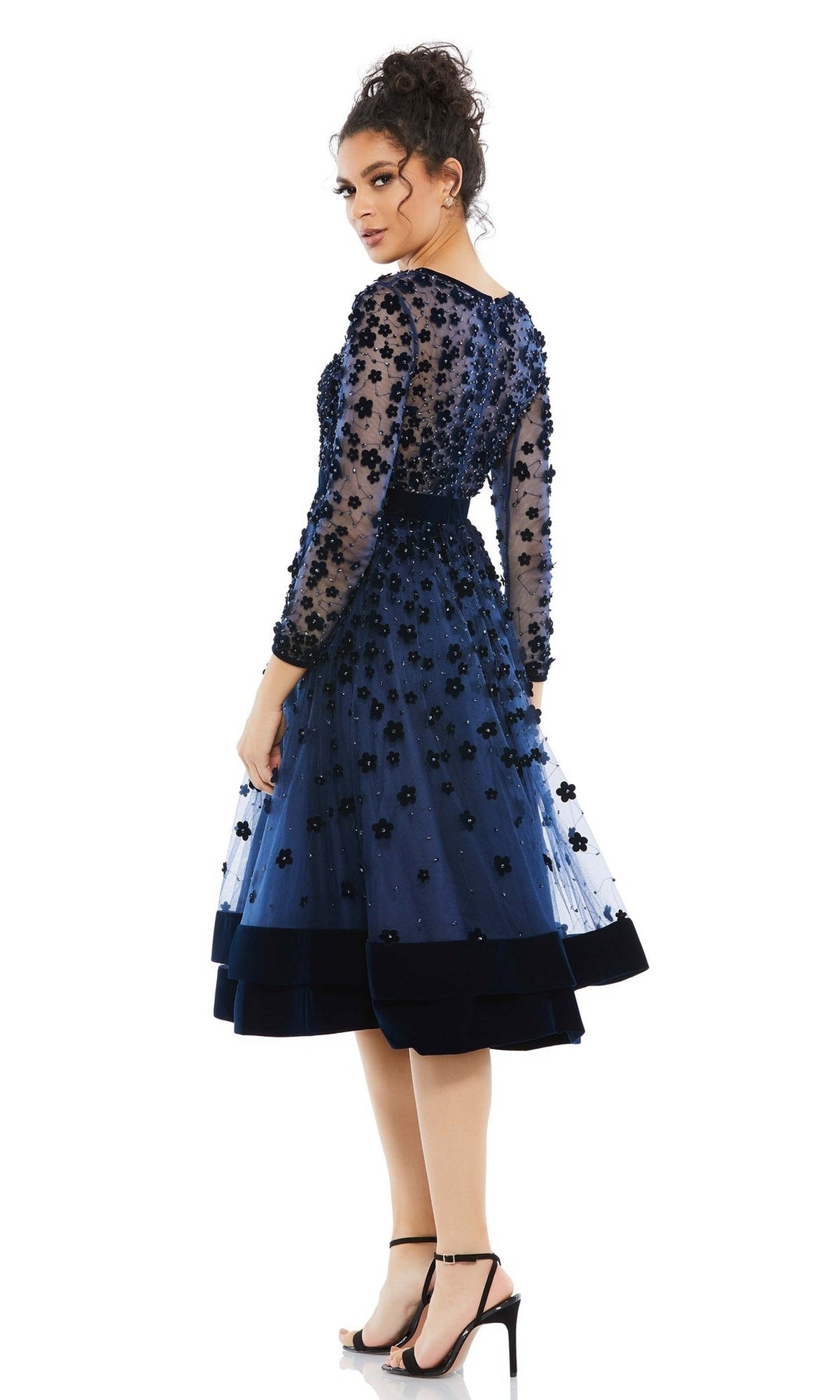 Woman wearing the Mac Duggal 67007 knee-length wedding-guest dress in navy blue with floral appliqué, beaded sheer long sleeves, and black high-heeled sandals, standing against a white background.